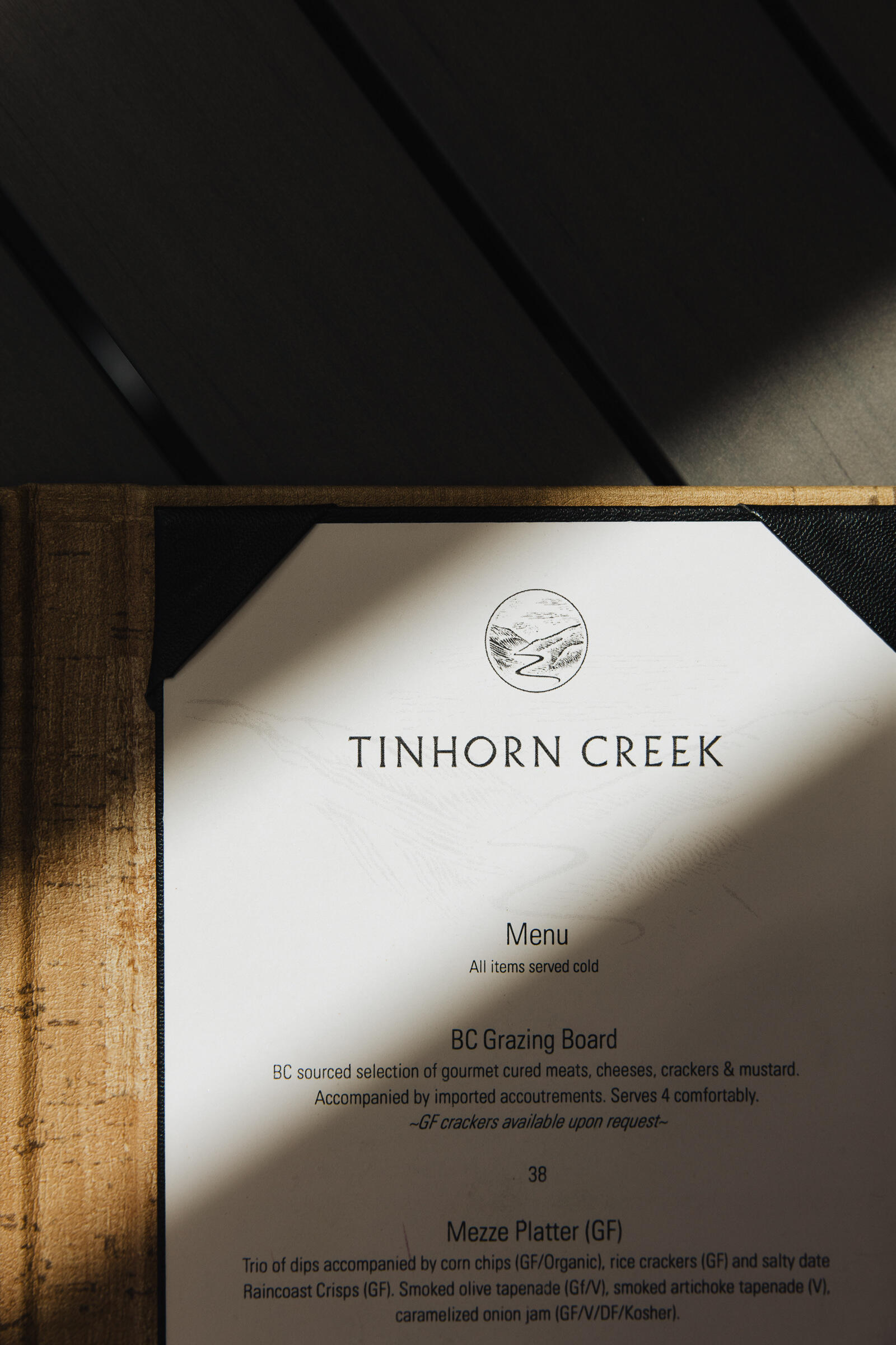 Close-up of a Tinhorn Creek menu on a wooden surface, partially in shadow. The menu lists items like a BC Grazing Board featuring meats, cheeses, crackers, mustard, and a Mezze Platter with tapenade and onion jam.