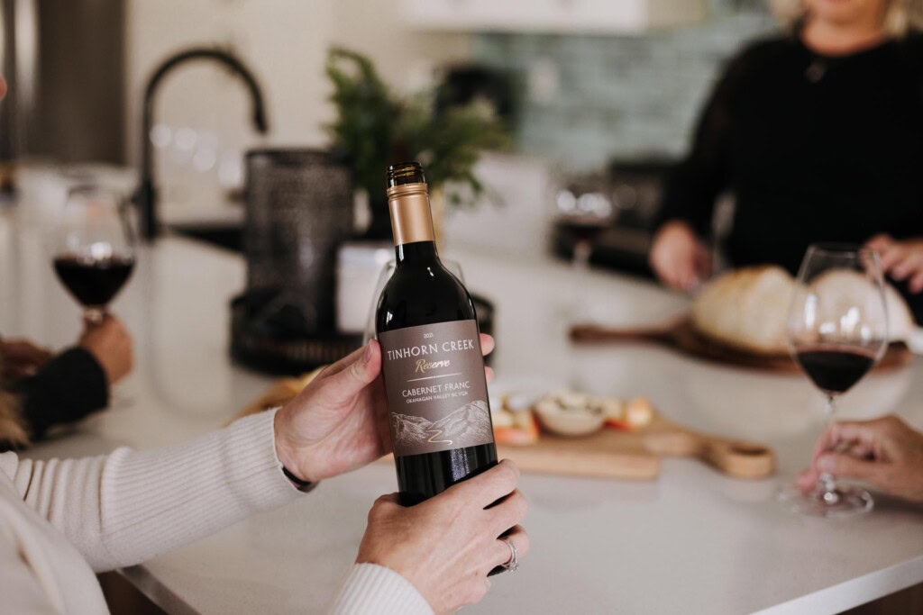 A person holds a bottle of Tinhorn Creek Cabernet Franc wine in a kitchen. Two filled wine glasses, a loaf of bread, and a wooden board with sliced apples and cheese are on the counter. Two other people are visible in the background.