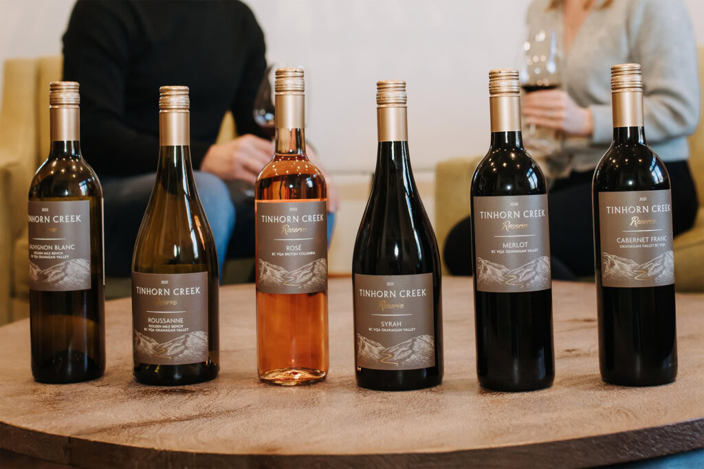 Six bottles of Tinhorn Creek wine are arranged on a wooden table. In the background, two people are seated having a conversation and holding wine glasses.