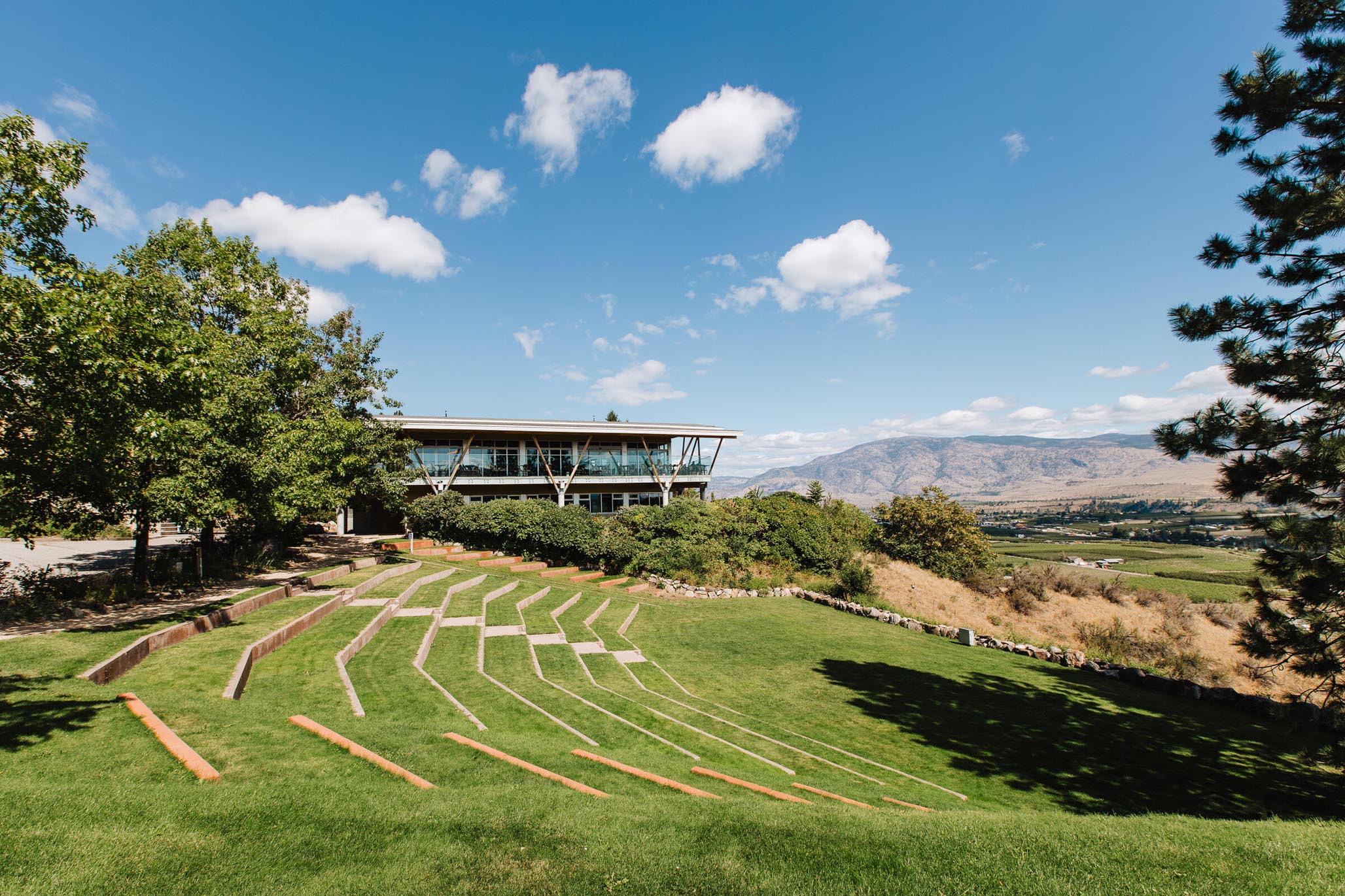 An outdoor amphitheater with grass terraces is situated near a modern building surrounded by trees. In the background, there are hills under a clear blue sky with scattered clouds.