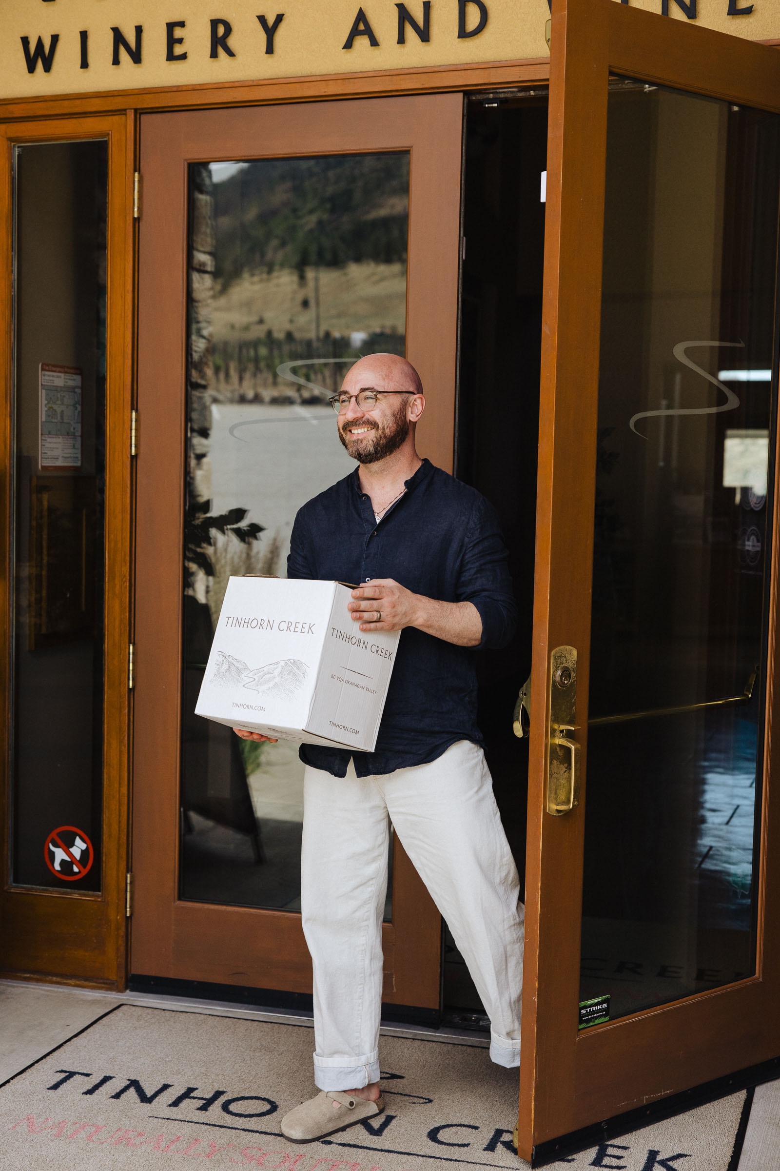 A man in a dark shirt and light pants steps out of a winery holding two boxes labeled "Tinhorn Creek." The entrance has glass doors, and winery signage is visible above.