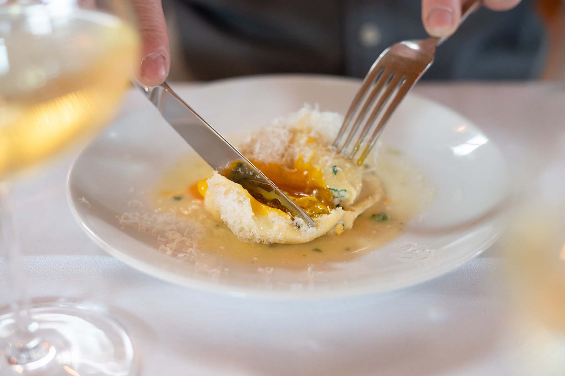 A person is slicing into a stuffed pasta dish with a fork and knife, revealing an egg yolk inside. The plate is garnished with grated cheese and herbs, and a glass of white wine is partially visible in the foreground.