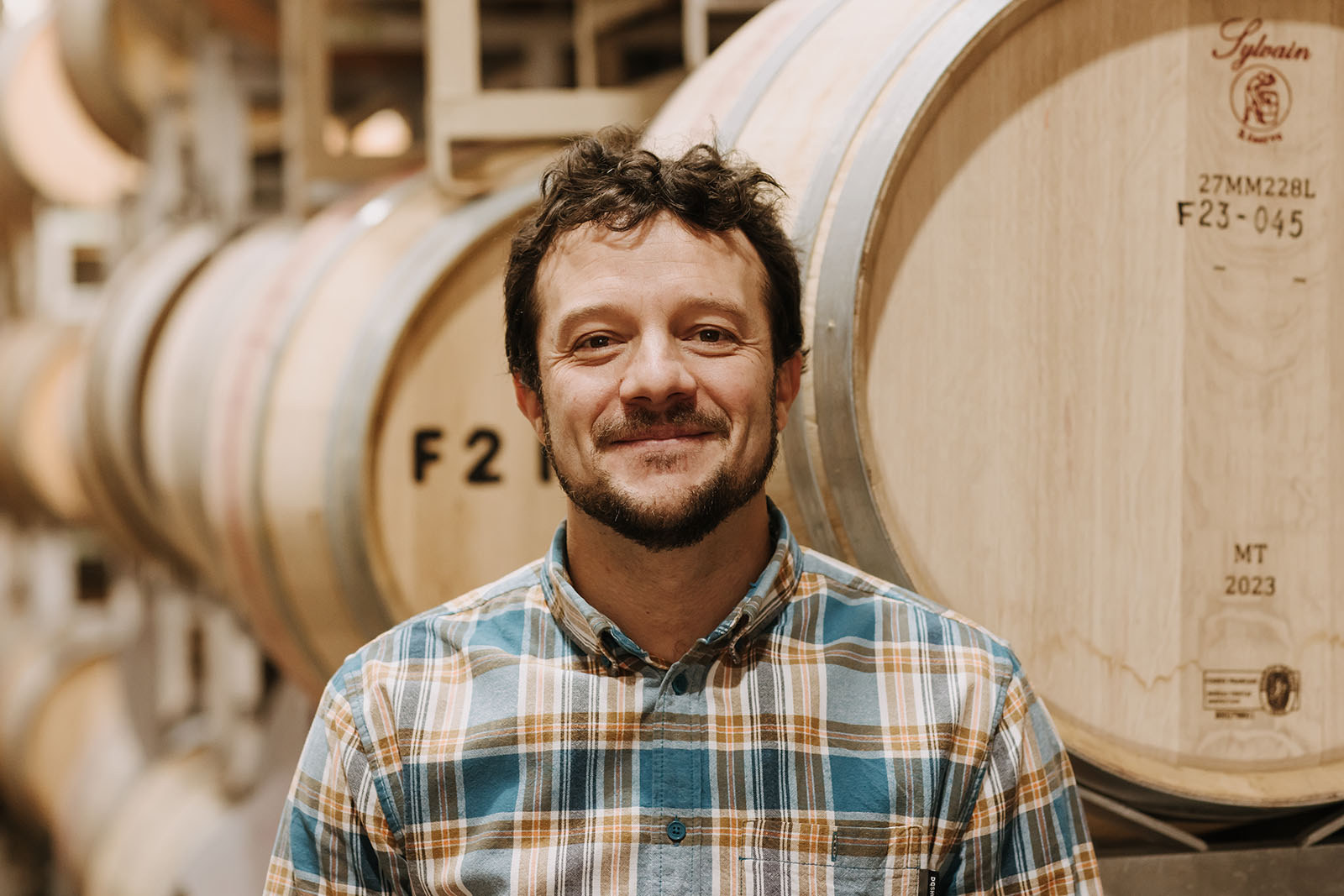 A man with curly hair and a beard stands smiling in front of stacked wine barrels. He is wearing a plaid shirt. The setting is a winery.