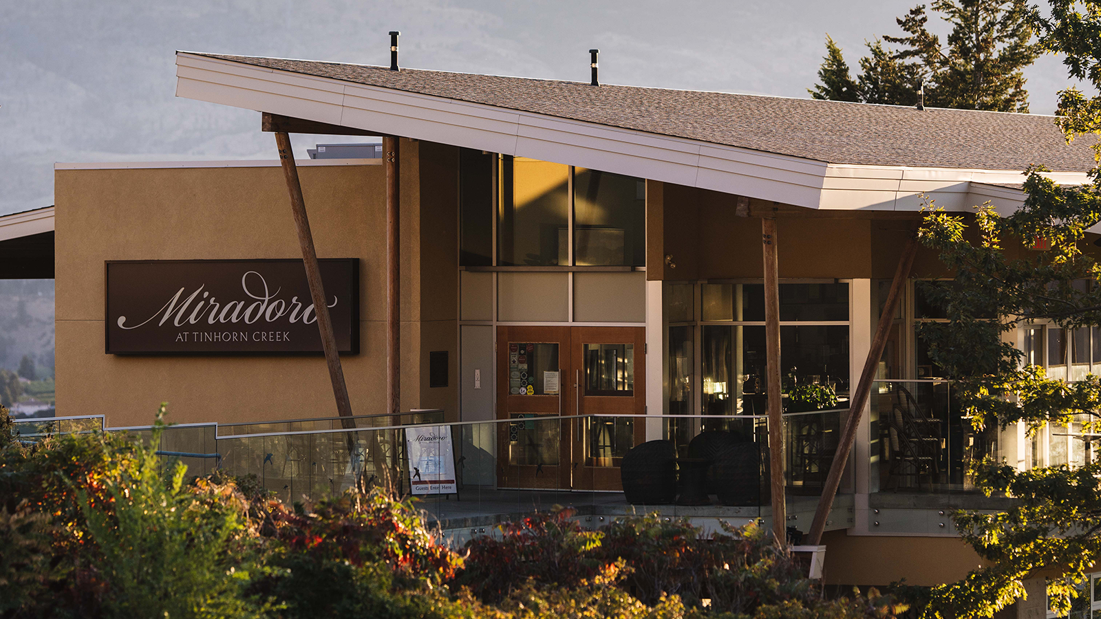 A modern building with large glass windows and a sloped roof sits among lush greenery, with a backdrop of rolling hills and a clear sky. The building features a sign that reads "Miradoro at Tinhorn Creek," indicating it is a restaurant. The exterior has earthy tones and wooden beams, complementing the surrounding landscape. The setting appears serene, illuminated by warm golden light, suggesting early morning or late afternoon.