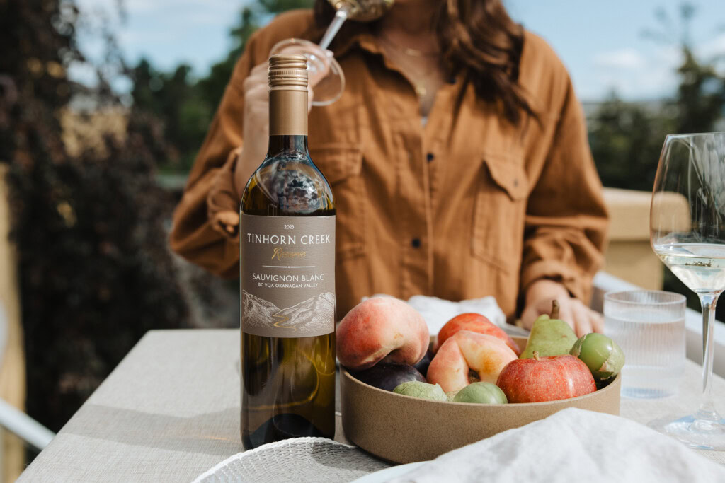 A bottle of Tinhorn Creek Sauvignon Blanc stands on a table next to a bowl of peaches and grapes. A person in a brown shirt is in the background, holding a wine glass.